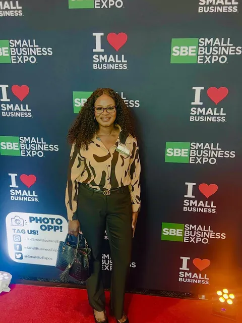 Mimi in a patterned blouse and dark trousers, handbag in hand, on the red carpet in front of the Small Business Expo backdrop in Dallas.